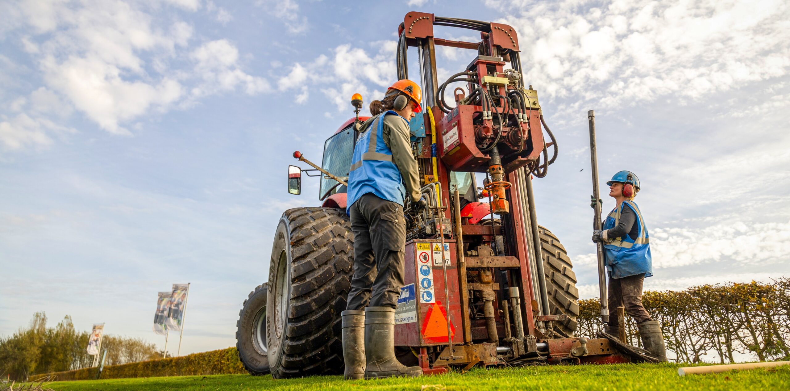 roter Traktor, blauer Himmel, auf Wiese, zwei Techniker bedienen Bohrgerät
