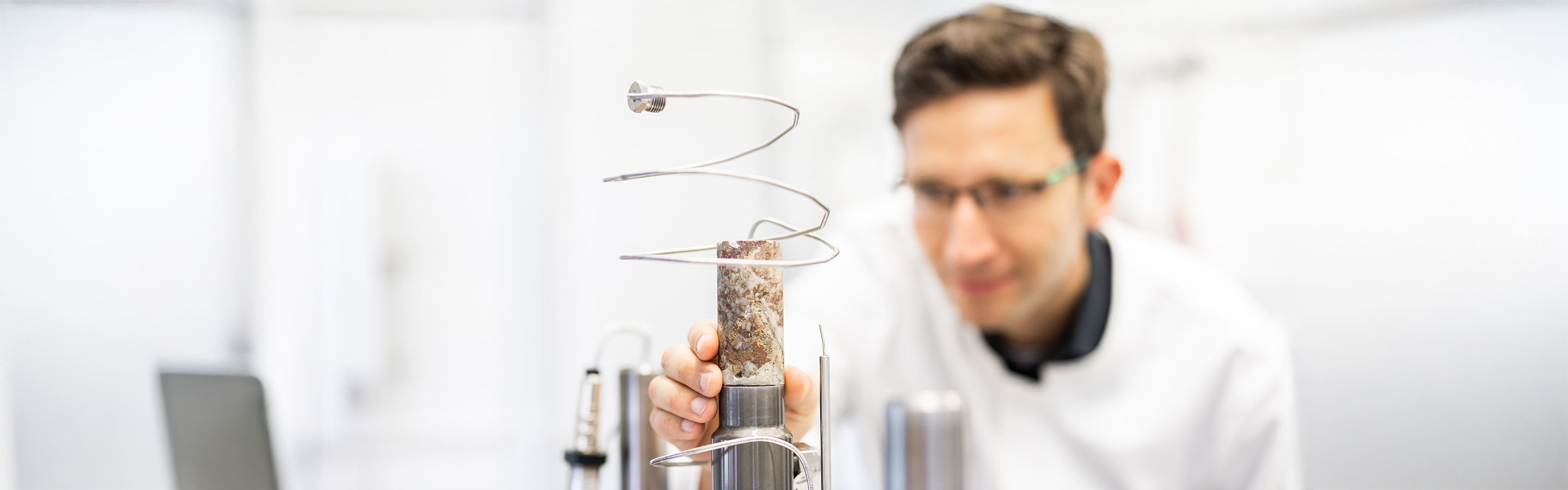 Mann im Labor hält kleine zylinderförmige Gesteinsprobe in der Hand (Vordergrund) um die eine metallerne Spirale befestigt ist.