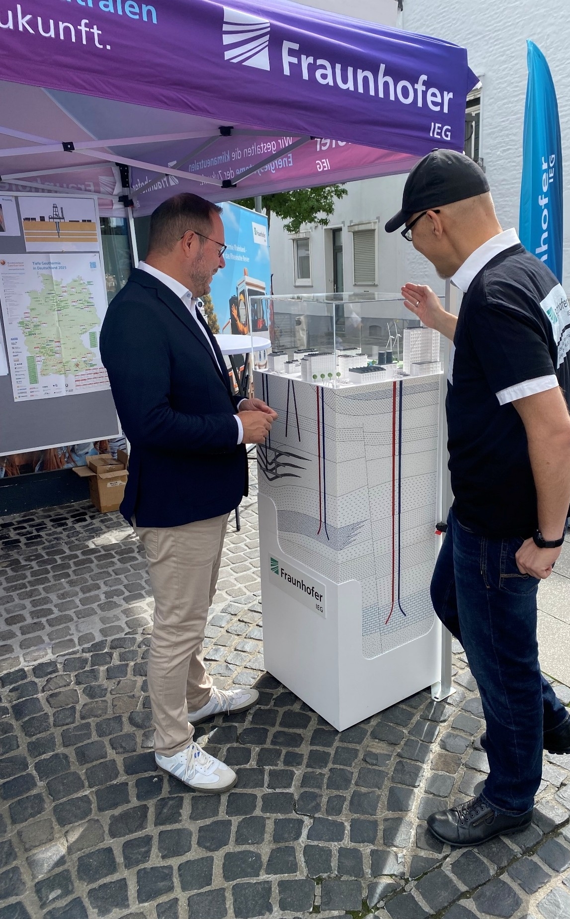 ALT-Text:  Unter einem bunten Pavillon mit der Aufschrift Fraunhofer IEG unterhalten sich zwei Männer. Beide betrachten dabei ein Modell, das Bohrungen in unterschiedliche Tiefen des Untergrundes darstellt. Auch auf diesem Modell ist das Logo des Fraunhofer IEGs sichtbar. Das Modell und der Pavillon stehen auf einer Straße, die mit Kopfsteinpflaster und Gehwegplatten gepflastert ist. Im Hintergrund ist eine Hauswand zu sehen.  