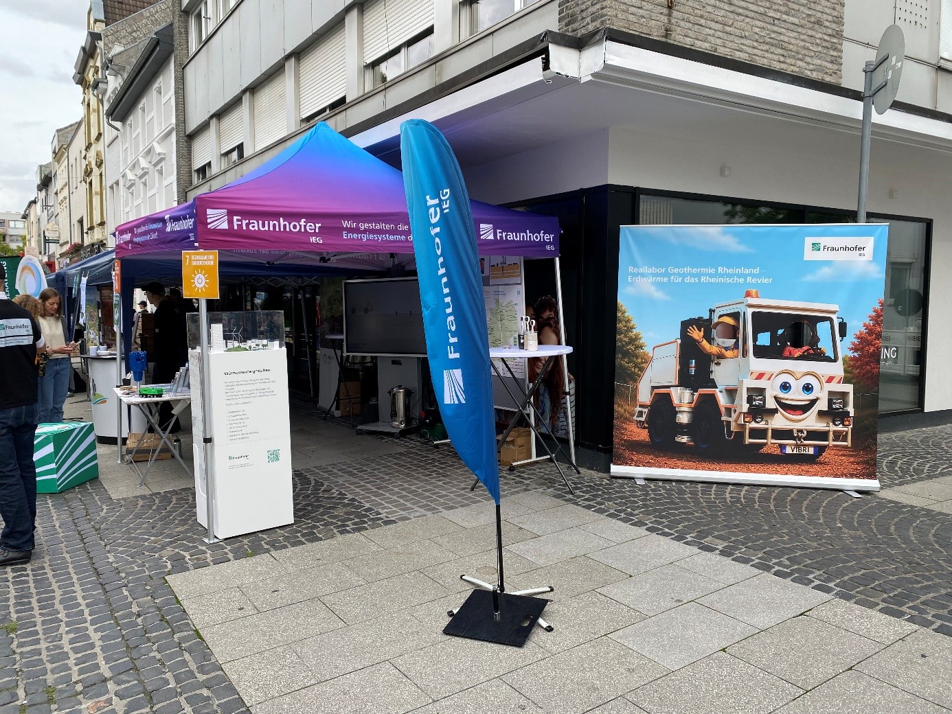 Informationsstand des Fraunhofer IEG in einer Fußgängerzone vor einem Geschäftsgebäude. Mehrere Pavillons stehen entlang des Gehwegs. Gleich als erster ein violett-blauer Pavillon mit dem Schriftzug „Fraunhofer IEG“ ergänzt durch eine blaue Fahne mit Fraunhofer-Logo. Vor dem Stand befinden sich Ausstellungselemente, ein Monitor und ein Modell zum Thema Erdwärme. Rechts zeigt ein großformatiges Guckloch-Banner den Text „Reallabor Geothermie Rheinland – Erdwärme für das Rheinische Revier“ mit der Abbildung eines Vibrotrucks.