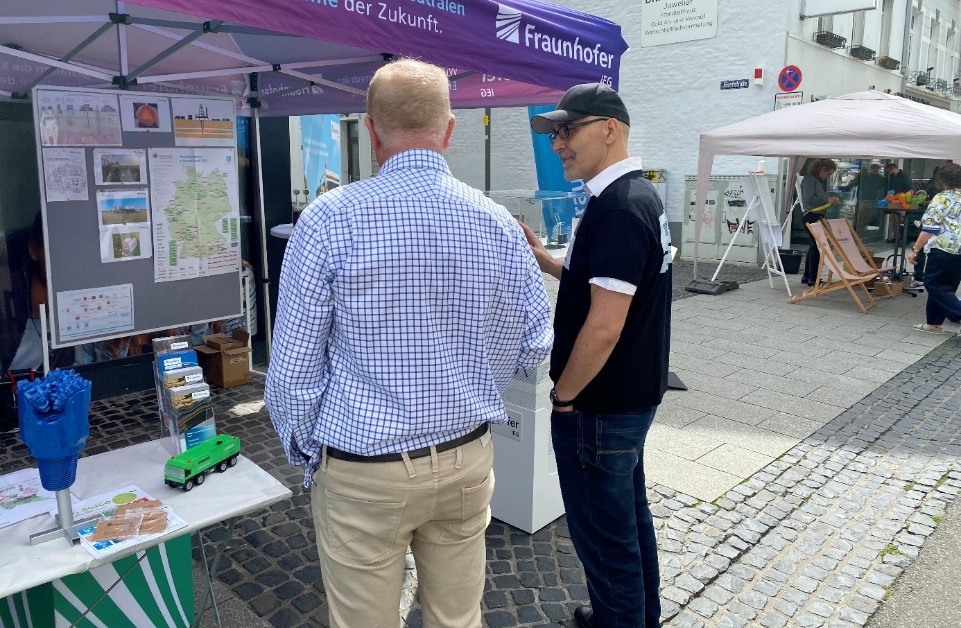 Zwei Personen stehen in einer Fußgängerzone unter einem Pavillon des Fraunhofer IEG mit der Aufschrift „Wir gestalten die klimaneutralen Energiesysteme der Zukunft“. Sie betrachten Ausstellungstafeln zum Thema Geothermie. Auf einem Tisch liegen Informationsmaterialien und ein Modellfahrzeug. Im Hintergrund sind weitere Pavillons, Passanten sowie die Fassade eines Geschäfts zu sehen. Die Szene zeigt eine öffentliche Informations- und Dialogveranstaltung im Stadtzentrum.